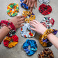 white and black hands reaching out to select a gold ankara scrunchie from a selection of 12 bright print options