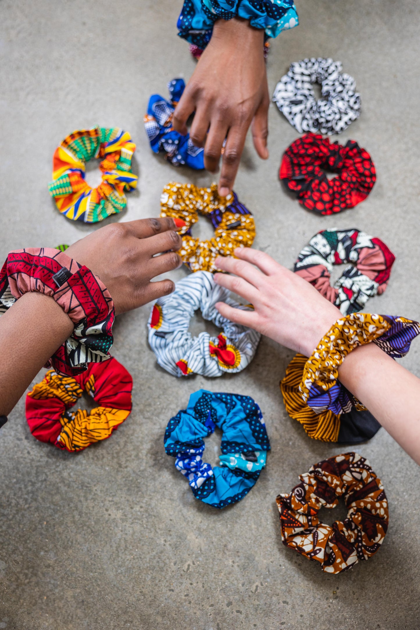 white and black hands reaching out to select a gold ankara scrunchie from a selection of 12 bright print options